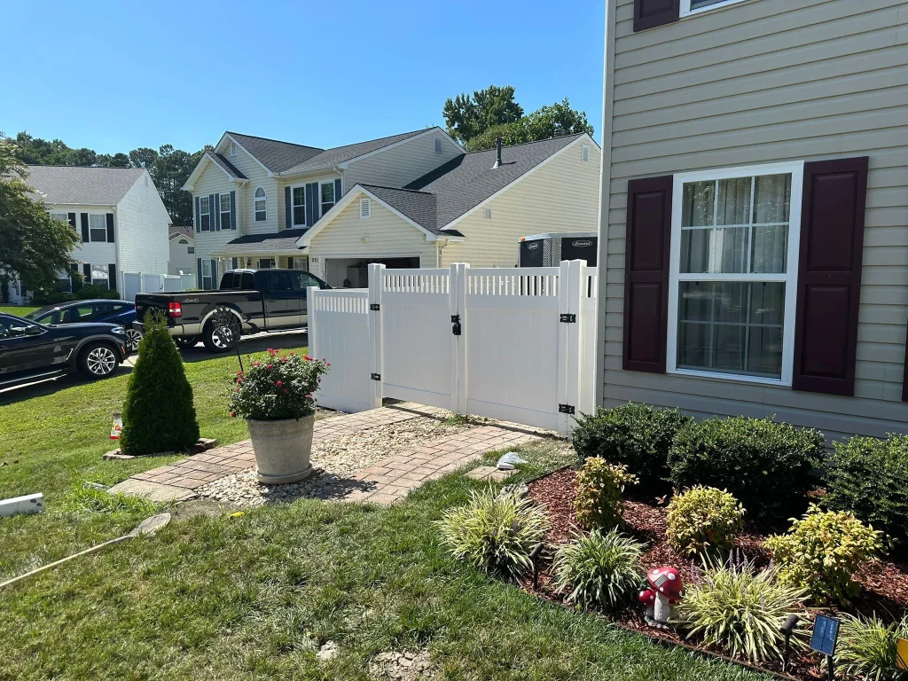 A white PVC double gate separates a landscaped front yard with a potted plant and shrubs from a driveway and other houses under a clear blue sky.